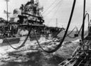 Its main deck piled with ammunition and fuel lines spanning a choppy sea, the Sacramento replenishes the aircraft carrier Ticonderoga (CVA-14) on Yankee Station. Note the six fuel hoses and two Burton rigs deployed as the AOE simultaneously supplies black oil, aviation fuel, ammunition, and stores. (U.S. Naval Institute)